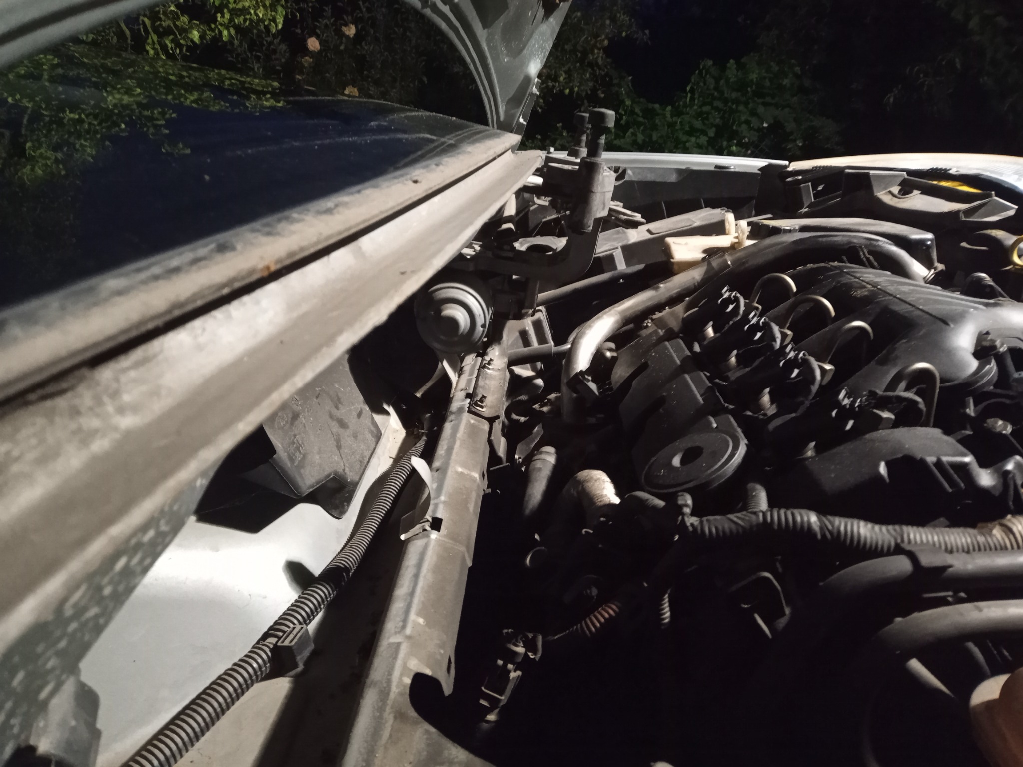 Otwarta maska samochodu, widoczne elementy silnika i instalacji elektrycznej, ciemne tło, słabe oświetlenie.