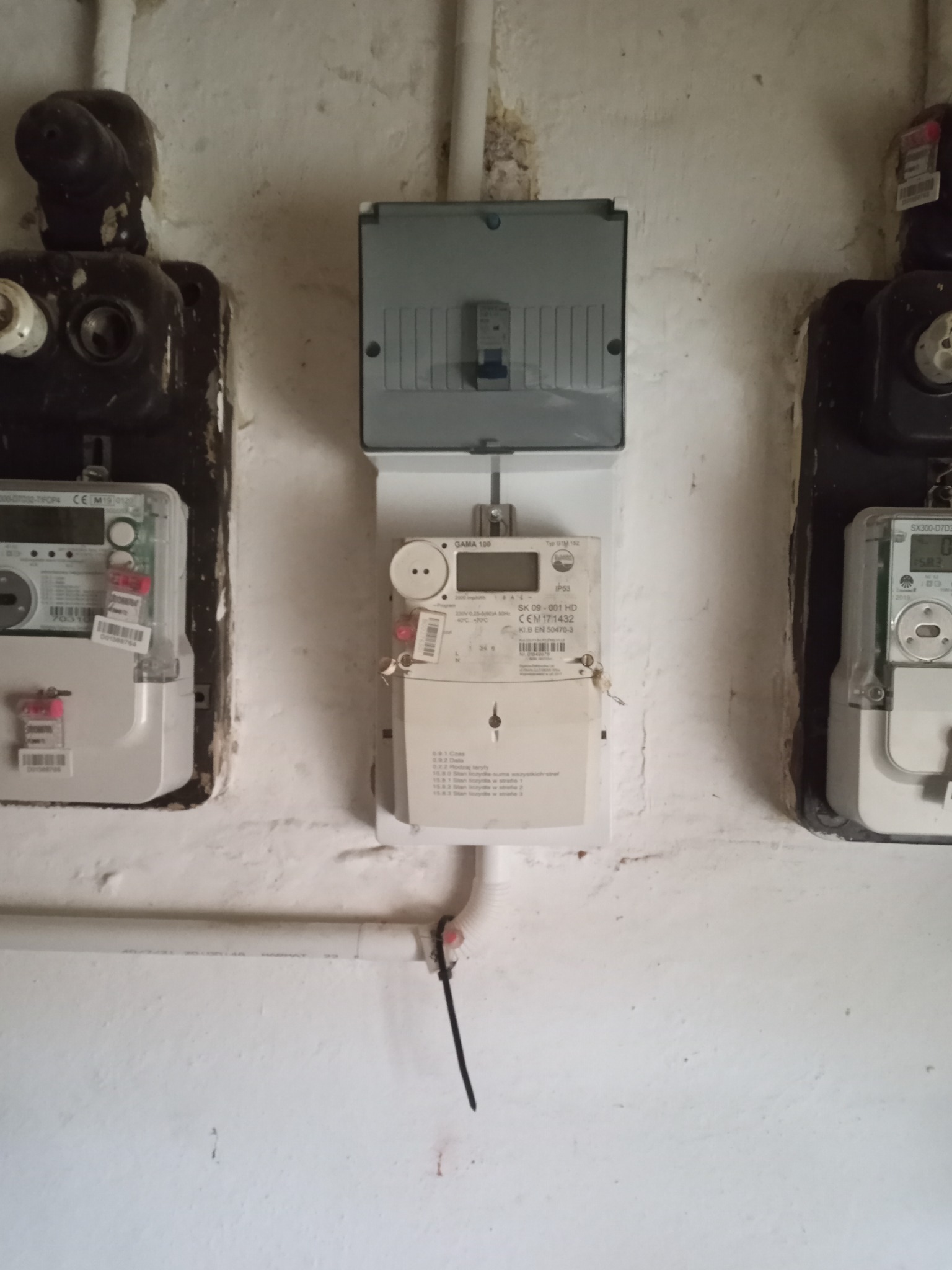 Przyłącze elektryczne: trzy liczniki energii elektrycznej zamontowane na pomalowanej na biało, starej ścianie, widoczne przewody i bezpieczniki.