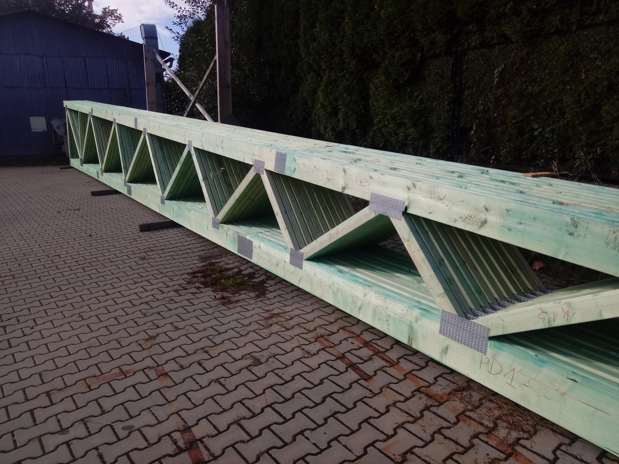 Impregnowana na zielono drewniana więźba dachowa z metalowymi łącznikami, leżąca na kostce brukowej przed budynkiem i żywopłotem.
