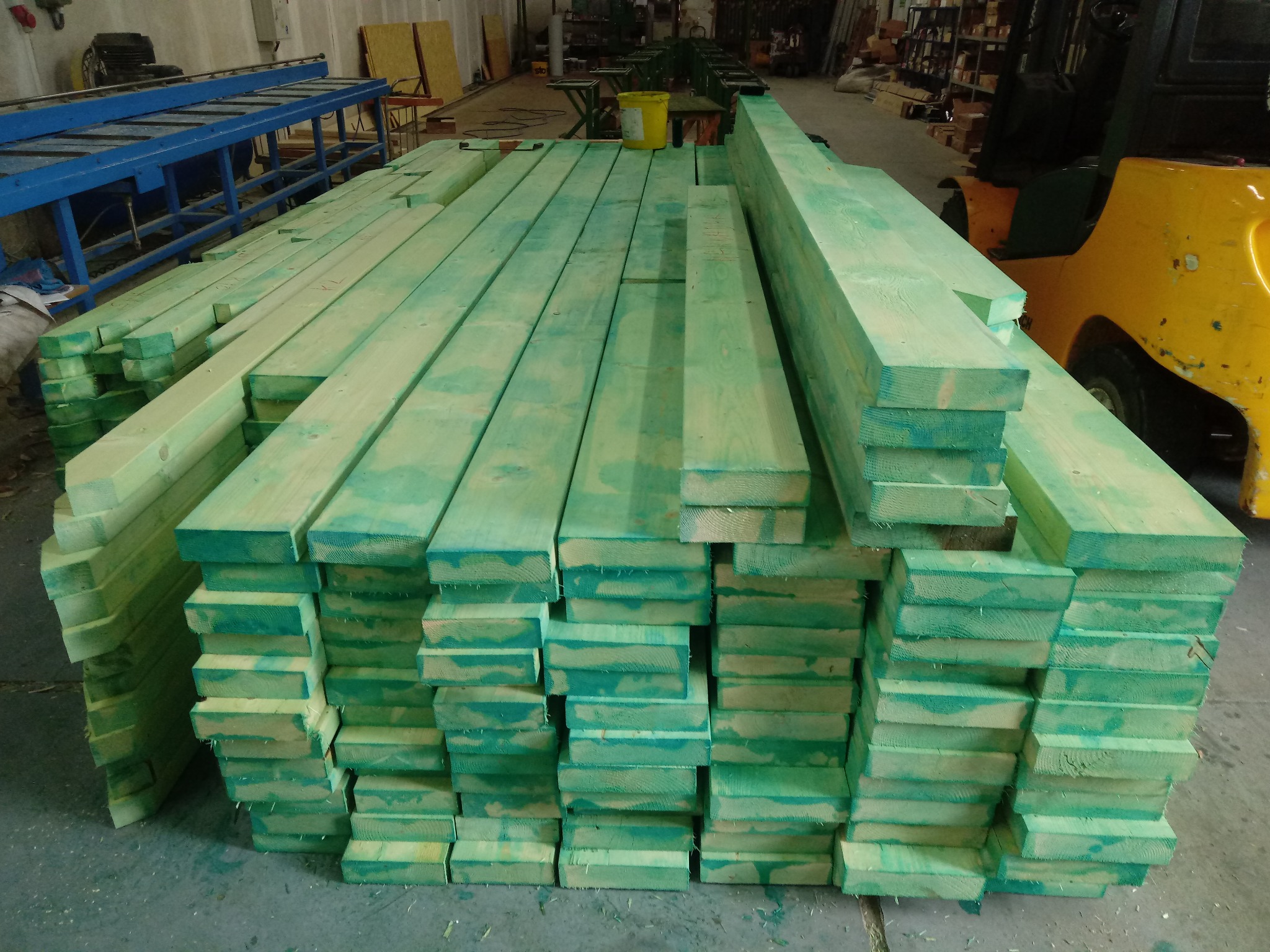 Stos impregnowanych na zielono kantówek ułożonych w magazynie, w tle wózek widłowy i niebieski stół transportowy.