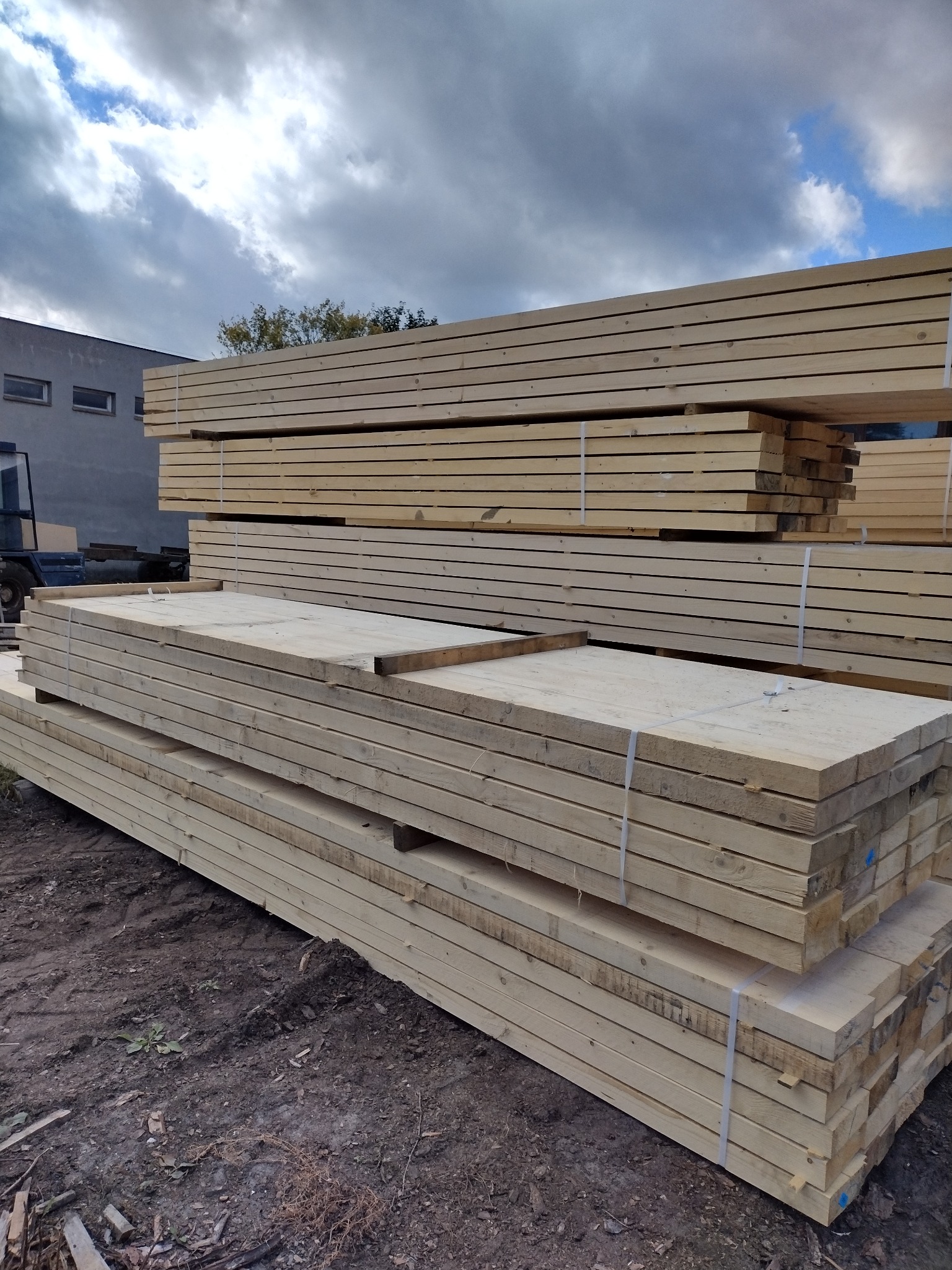 Stosy świeżego, jasnego drewna budowlanego, zabezpieczone białymi taśmami, ułożone na ziemi z widocznym budynkiem w tle pod pochmurnym niebem.