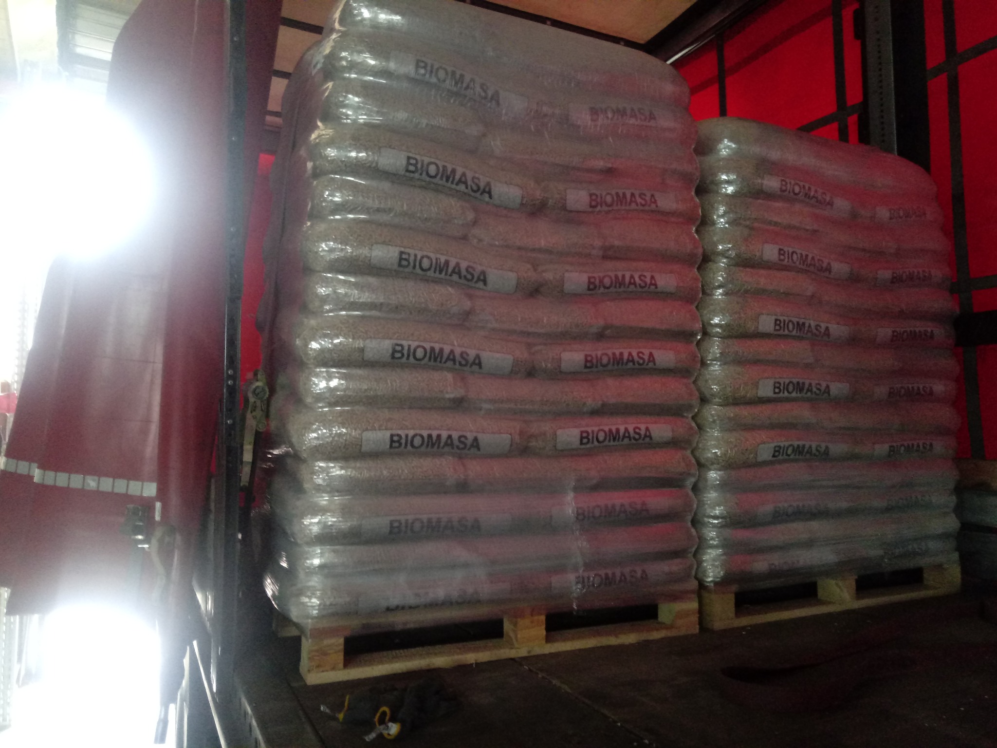 Załadunek palet z workami biomasy, opakowanymi w folię, wewnątrz naczepy ciężarówki z czerwonymi ścianami.