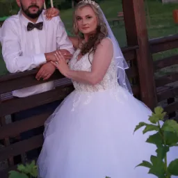 Para młoda w plenerze: Panna młoda w koronkowej sukni i welonie, Pan młody w białej koszuli i muszce, opierają się o drewnianą konstrukcję, widoczne obrączki.