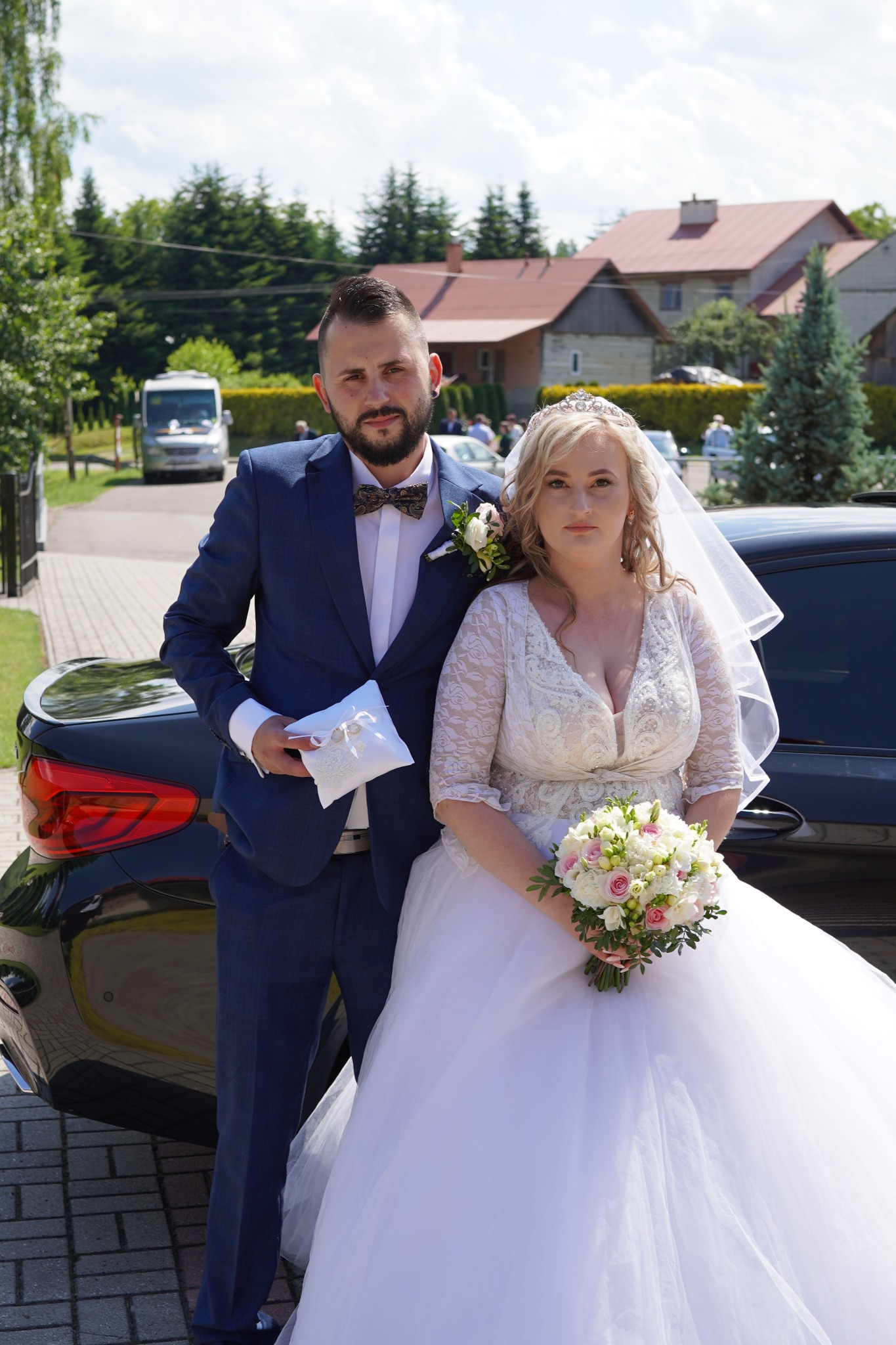 Para młoda w strojach ślubnych pozuje przy czarnym samochodzie. Pan młody w granatowym garniturze i muszce w drobny wzór trzyma poduszkę na obrączki. Panna młoda w koronkowej sukni z bukietem...