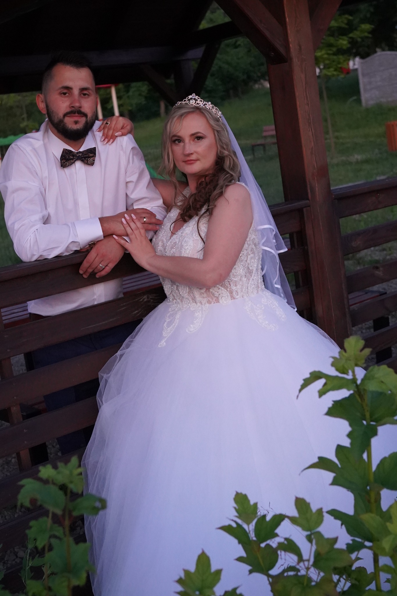 Para młoda w plenerze: Panna młoda w koronkowej sukni i welonie, Pan młody w białej koszuli i muszce, opierają się o drewnianą konstrukcję, widoczne obrączki.