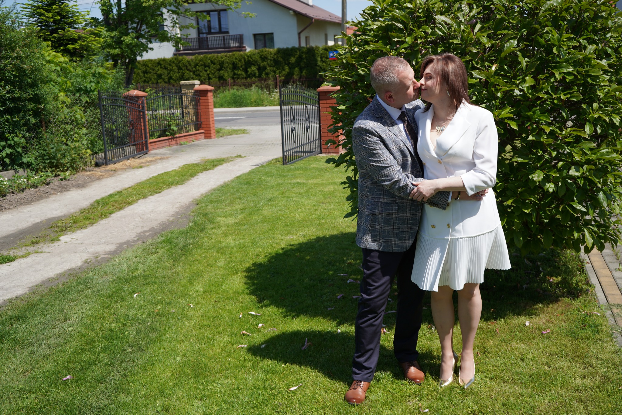 Elegancko ubrana para w średnim wieku całuje się na tle zieleni, w tle brama i dom jednorodzinny.