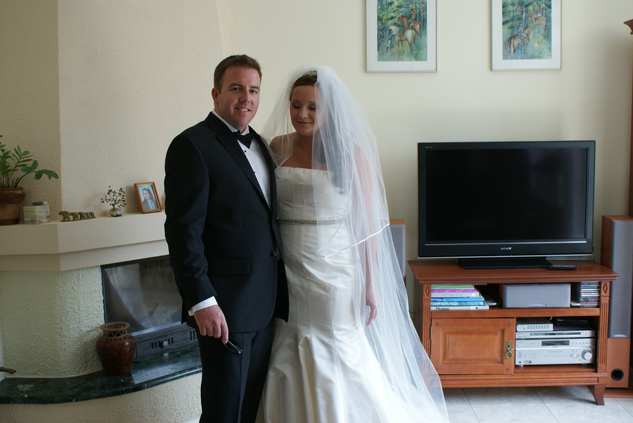 Para młoda w strojach ślubnych pozuje w salonie z kominkiem, dekoracjami i telewizorem.