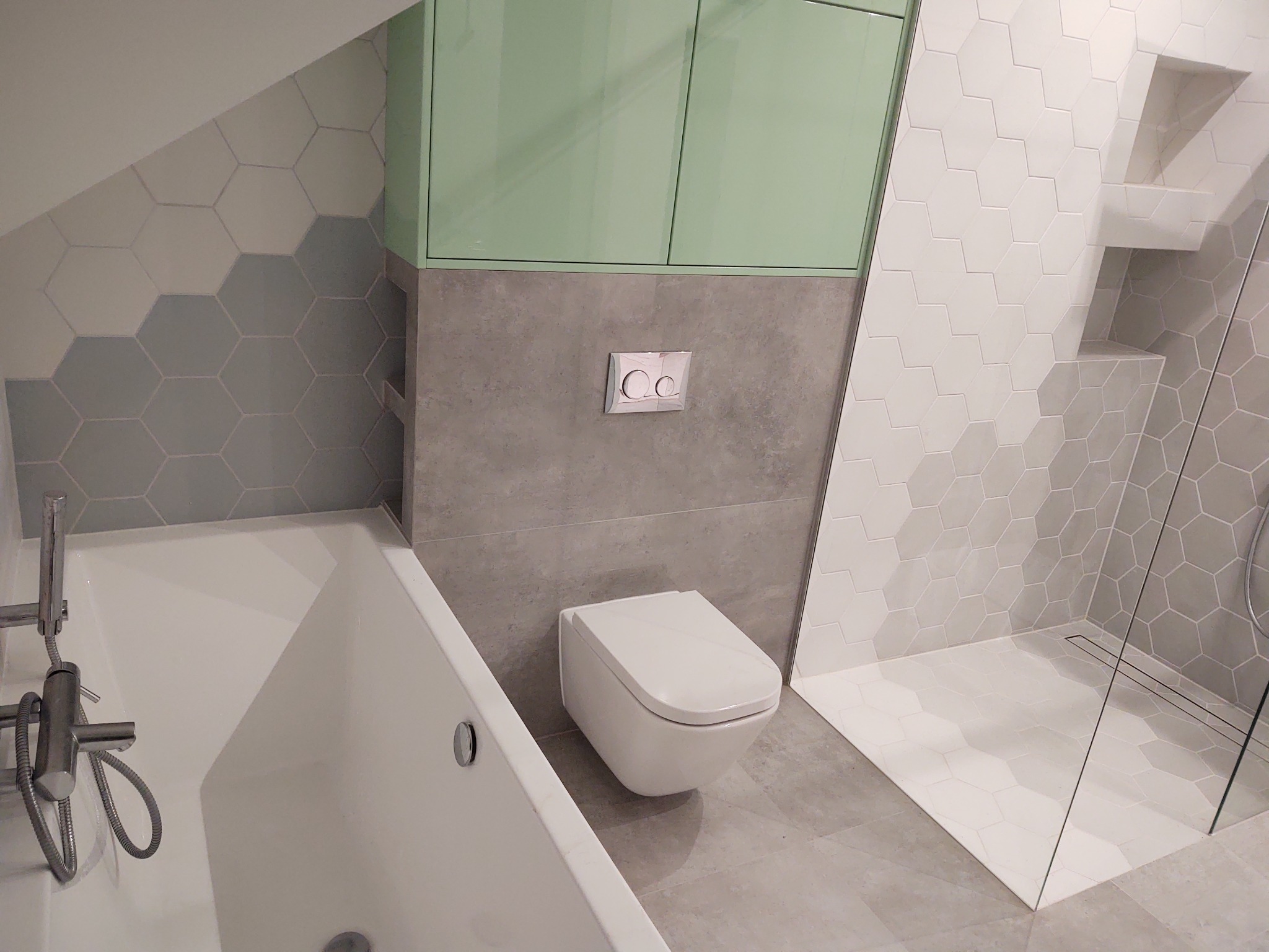 Nowoczesna łazienka z wanną, podwieszaną toaletą, kabiną prysznicową i heksagonalnymi płytkami w odcieniach szarości i bieli, uzupełniona szafką w kolorze miętowym.