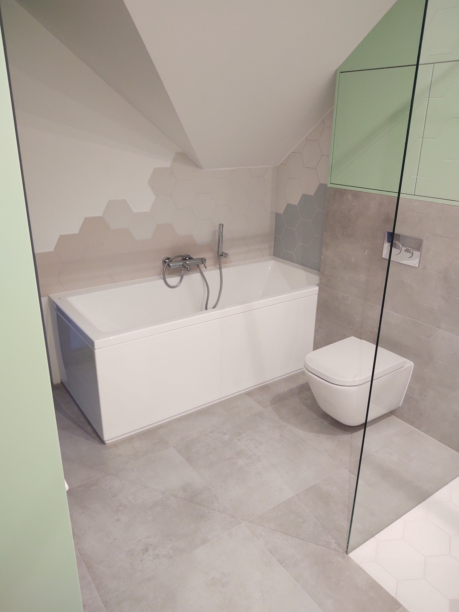 Nowoczesna łazienka na poddaszu z białą wanną, podwieszaną toaletą, szarą glazurą w geometryczne wzory i zabudową w kolorze pistacjowym.