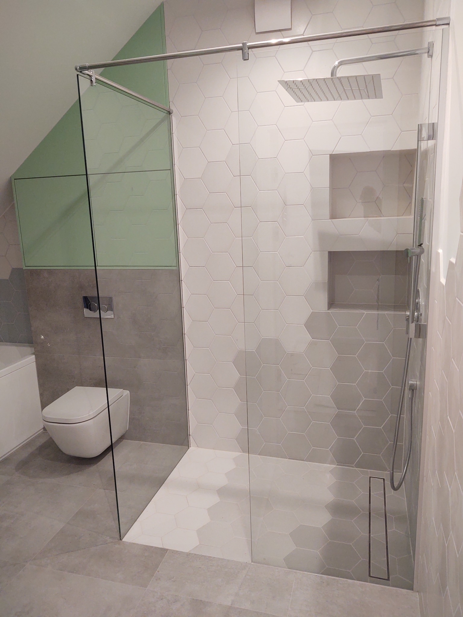 Nowoczesna łazienka z kabiną prysznicową typu walk-in, heksagonalne płytki w odcieniach szarości i bieli, minimalistyczna toaleta podwieszana i zabudowa w kolorze pistacjowym.