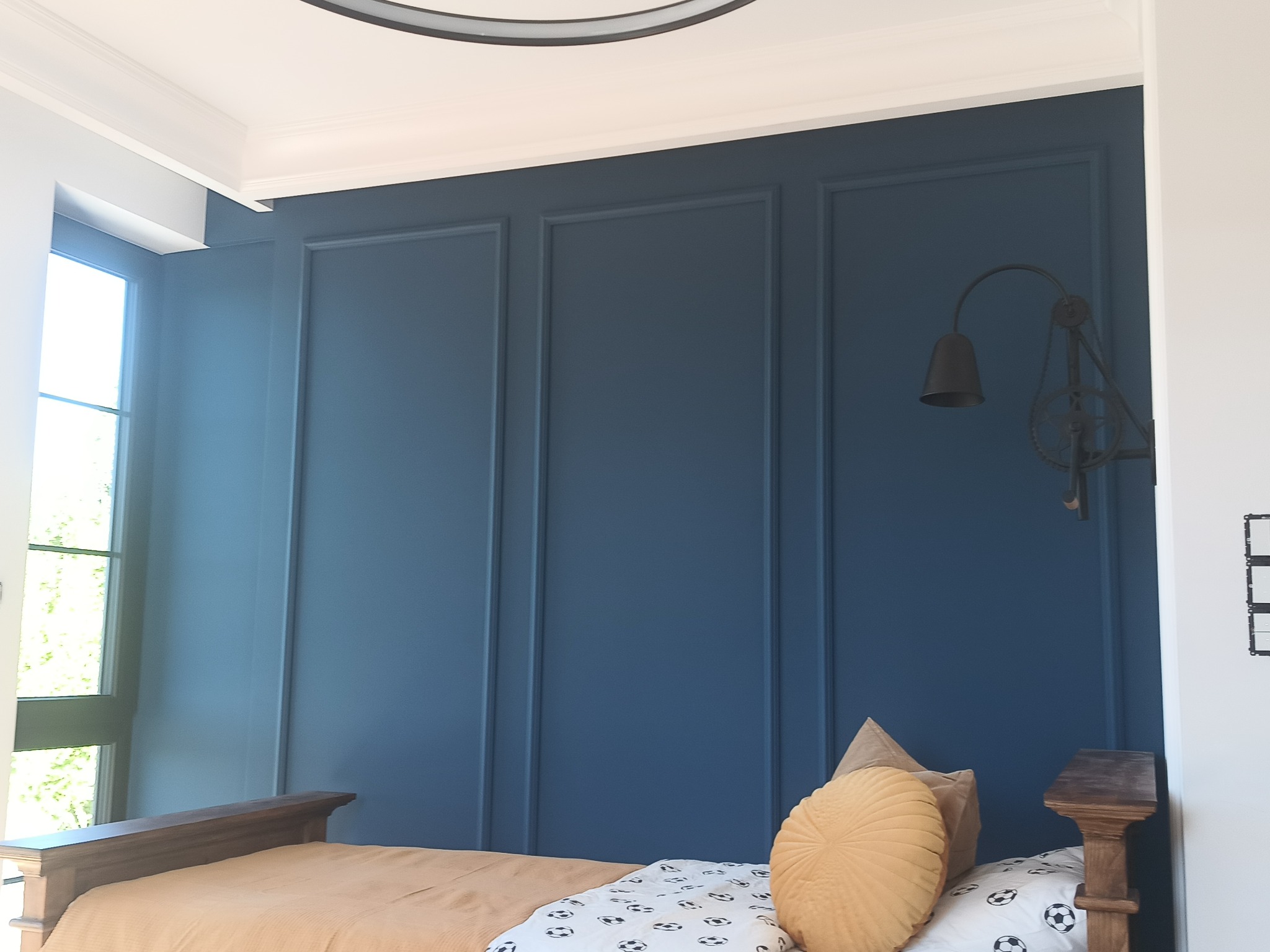 Niebieska ściana z dekoracyjnymi panelami w sypialni, oświetlona lampą w stylu industrialnym. Widoczne łóżko z beżową narzutą i poduszkami.