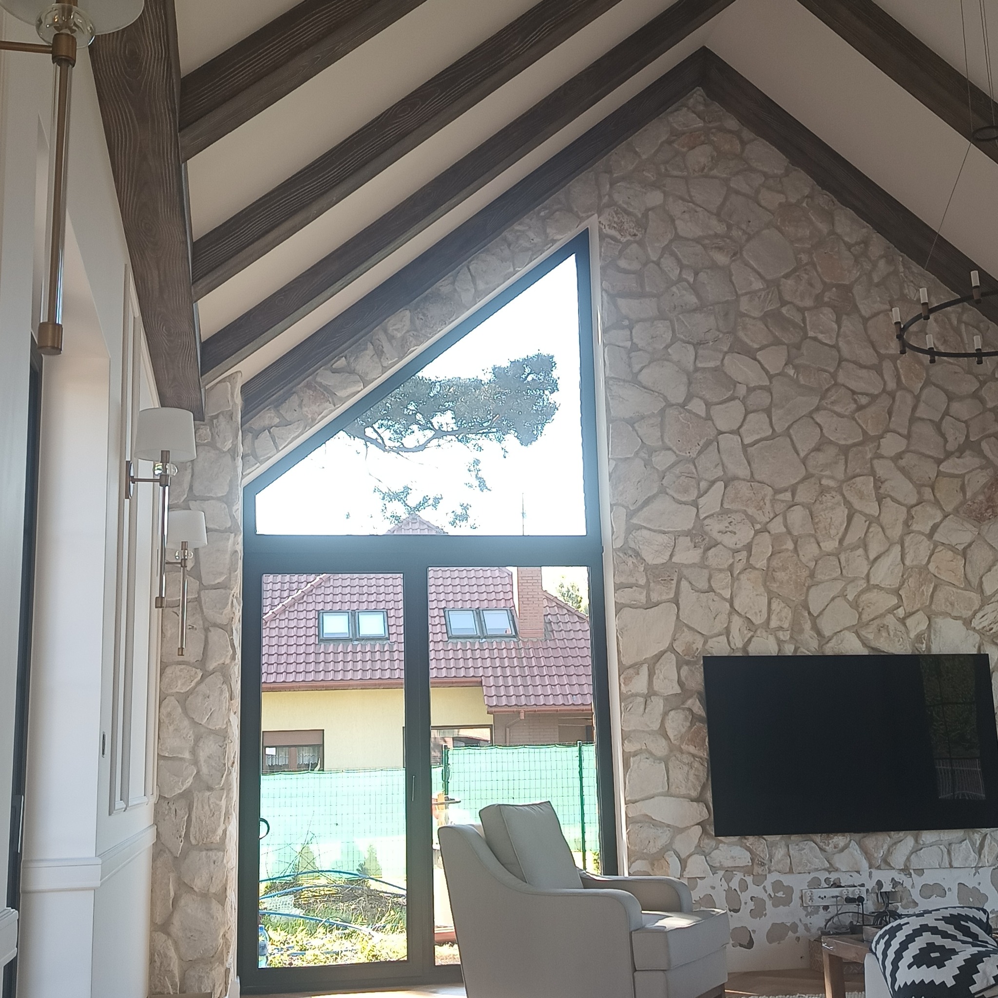 Salon z wysokim stropem, kamienną ścianą i dużym oknem trójkątnym. Widoczne drewniane belki stropowe i nowoczesne oświetlenie. W tle widok na ogród.