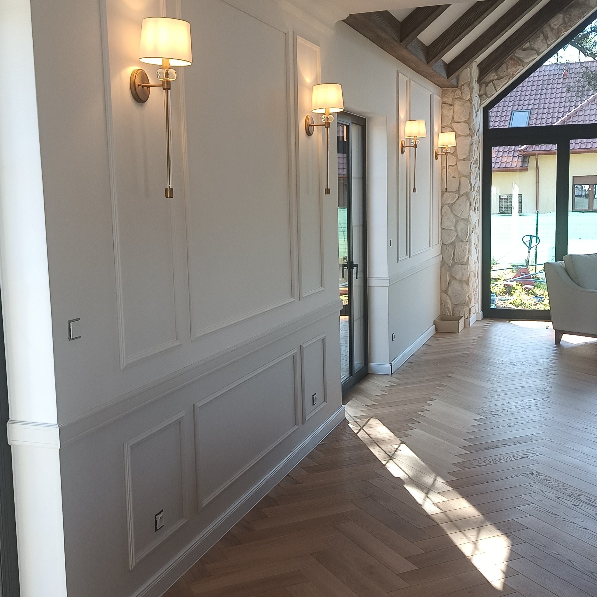 Eleganckie wnętrze z parkietem w jodełkę, ozdobnymi panelami ściennymi i złotymi kinkietami. Widok na ogród przez przeszklone drzwi.