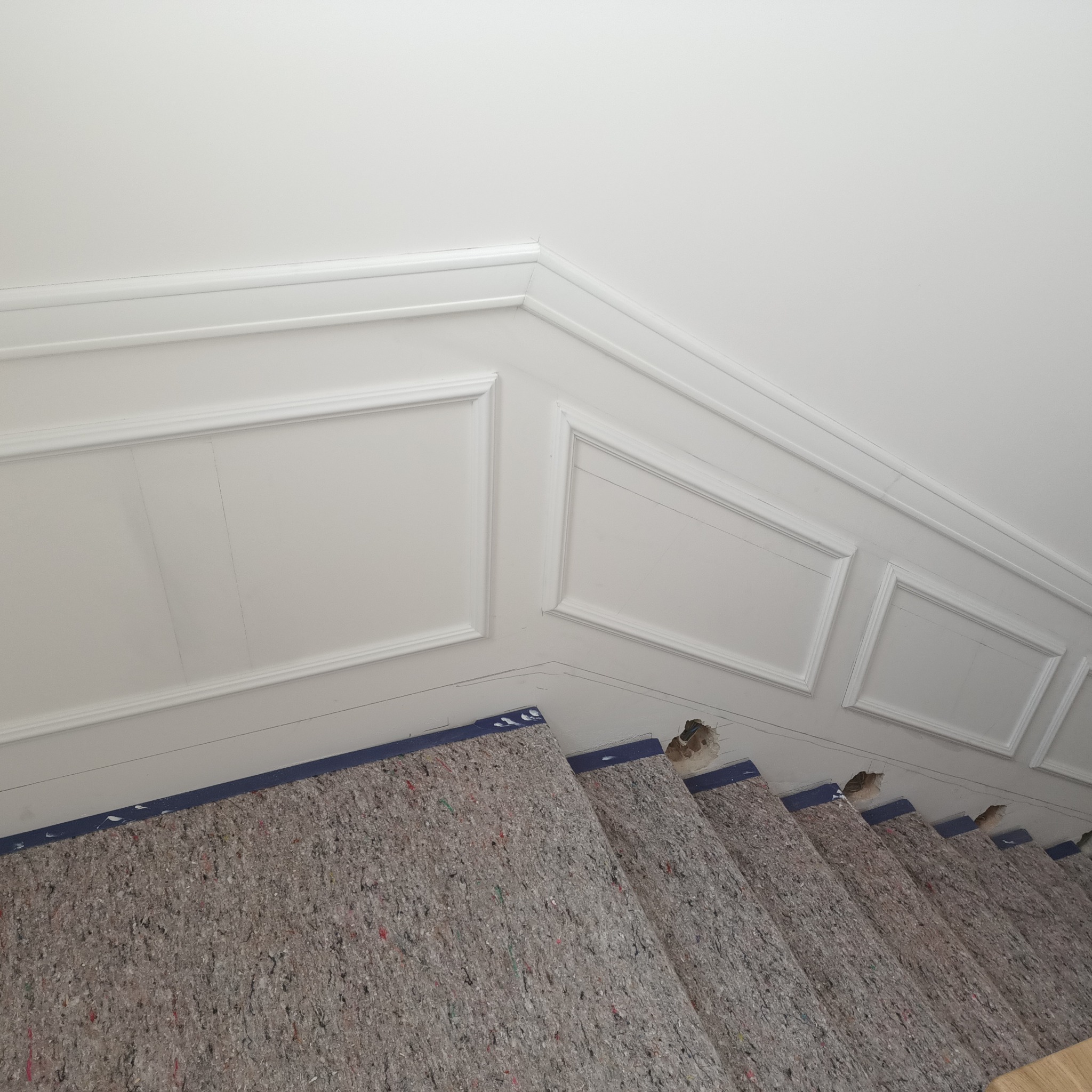 Białe, ozdobne panele ścienne z listwami i zabezpieczone schody z szarą wykładziną ochronną, widok z góry, podczas prac wykończeniowych.