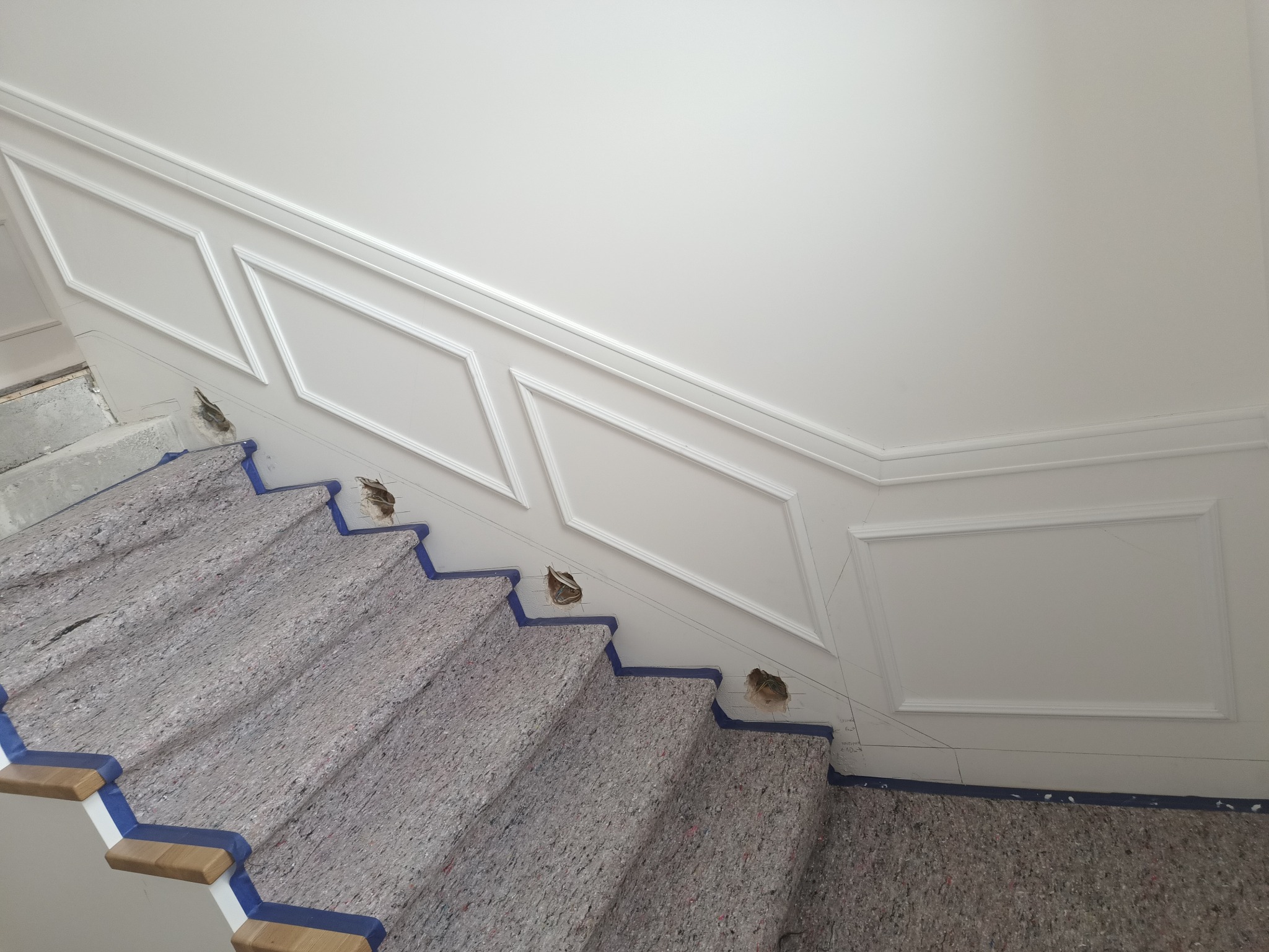 Białe, ozdobne listwy ścienne wzdłuż schodów zabezpieczonych folią malarską. Widoczne otwory w ścianie. Górne oświetlenie. Ukończony etap prac wykończeniowych.