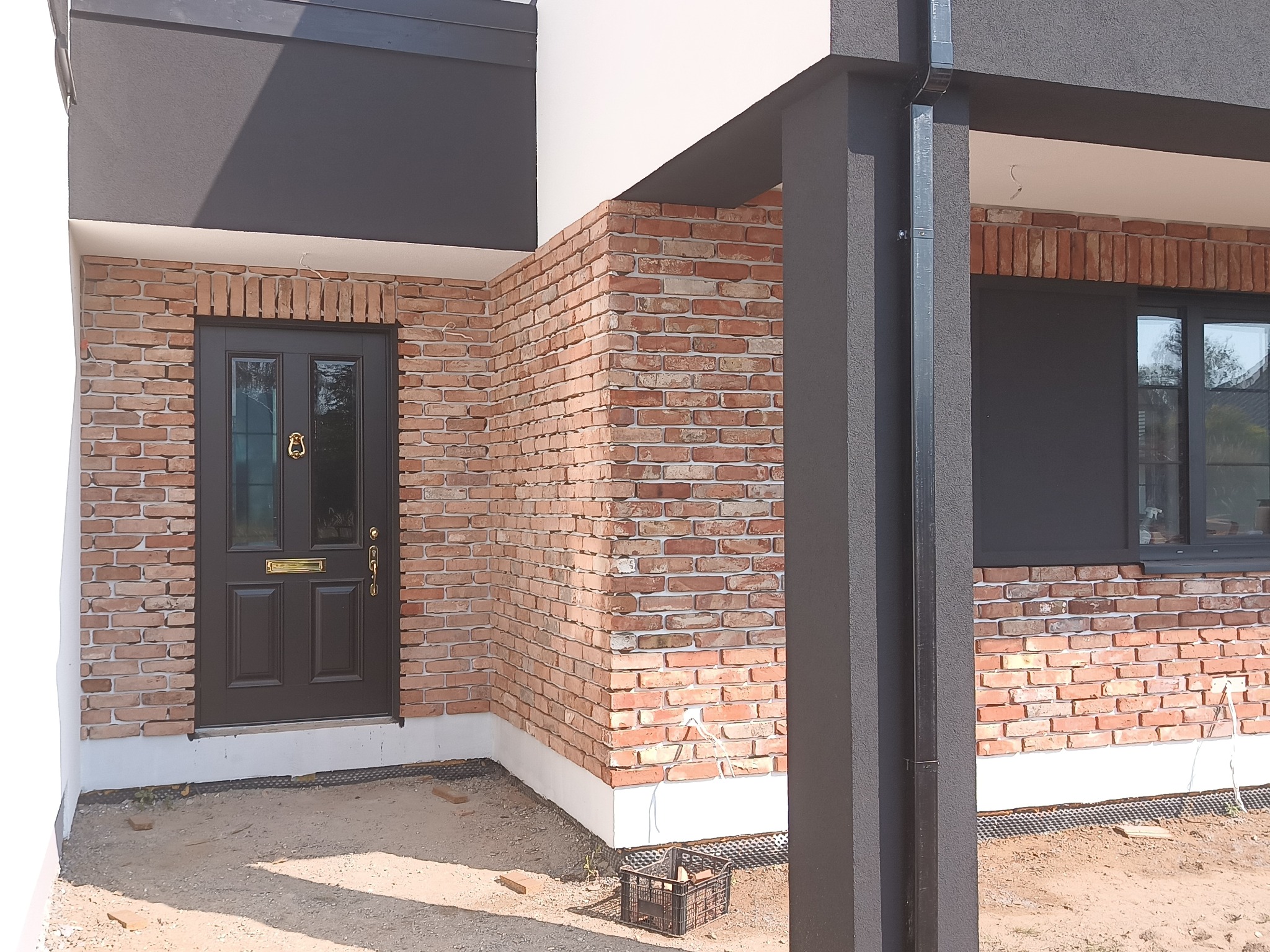 Elewacja budynku z cegły klinkierowej i ciemnymi elementami wykończeniowymi, widoczna stolarka okienna i drzwiowa, oraz fragment terenu przed budynkiem.