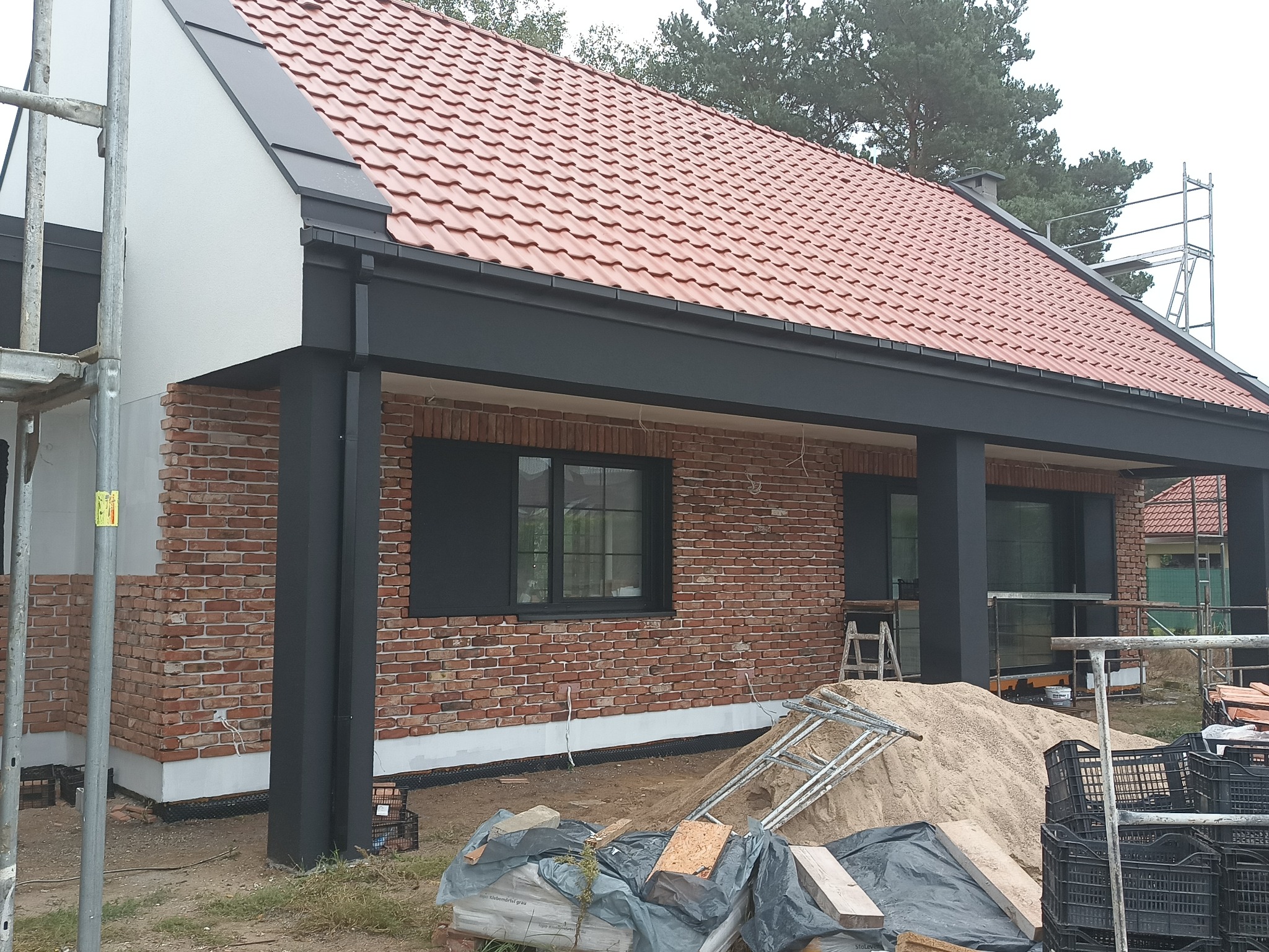 Dom z czerwoną dachówką i elewacją z cegły klinkierowej, czarne ramy okienne i kolumny, widoczne rusztowania i materiały budowlane.