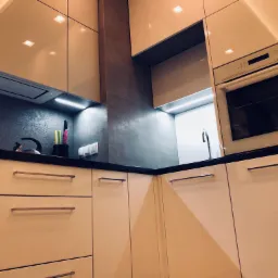 Beżowa, lakierowana kuchnia z czarnym blatem, oświetleniem LED podszafkowym i sprzętem AGD w zabudowie. Widoczne detale: czajnik, pojemnik na przybory kuchenne, gładkie fronty szuflad i szafek.