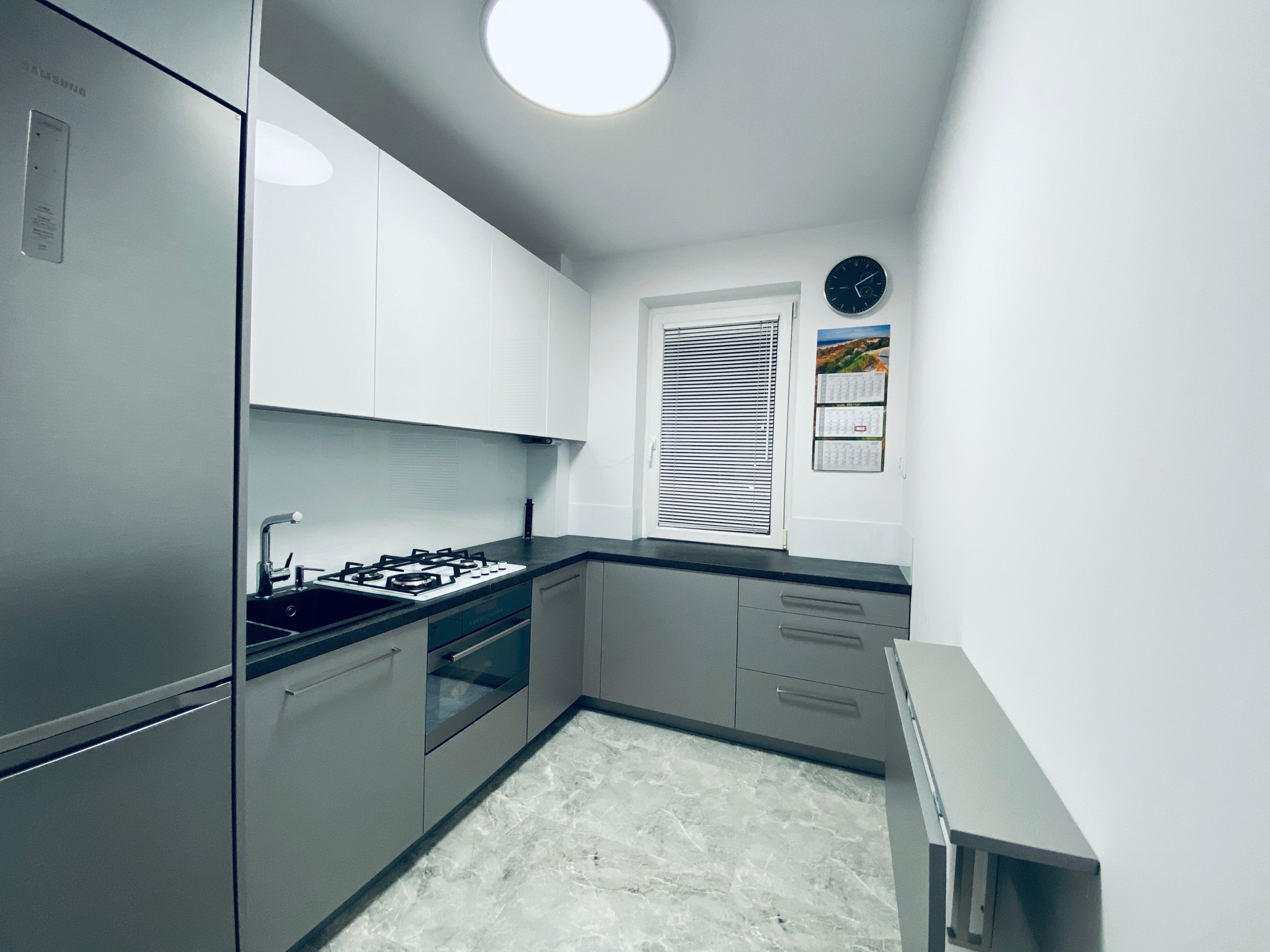 Mała kuchnia w bloku z szarymi frontami (dolne szafki) i białymi (górne szafki), czarnym blatem, płytą gazową, piekarnikiem i szarym marmurkowym wzorem na podłodze. Na ścianie widoczny zegar...