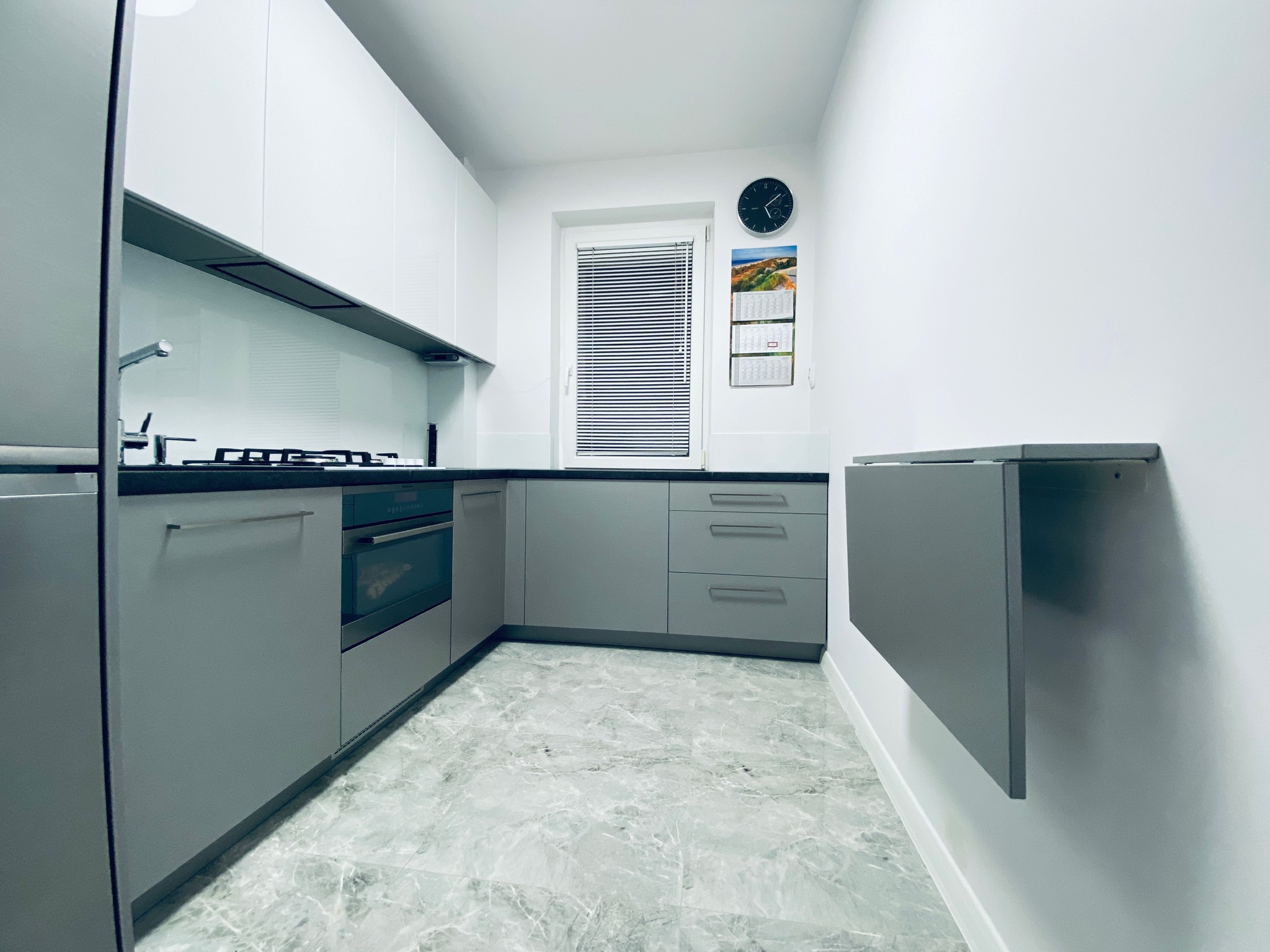 Nowoczesna, minimalistyczna kuchnia w odcieniach szarości i bieli, z marmurową podłogą i składanym blatem przy ścianie, oświetlona neutralnym światłem.