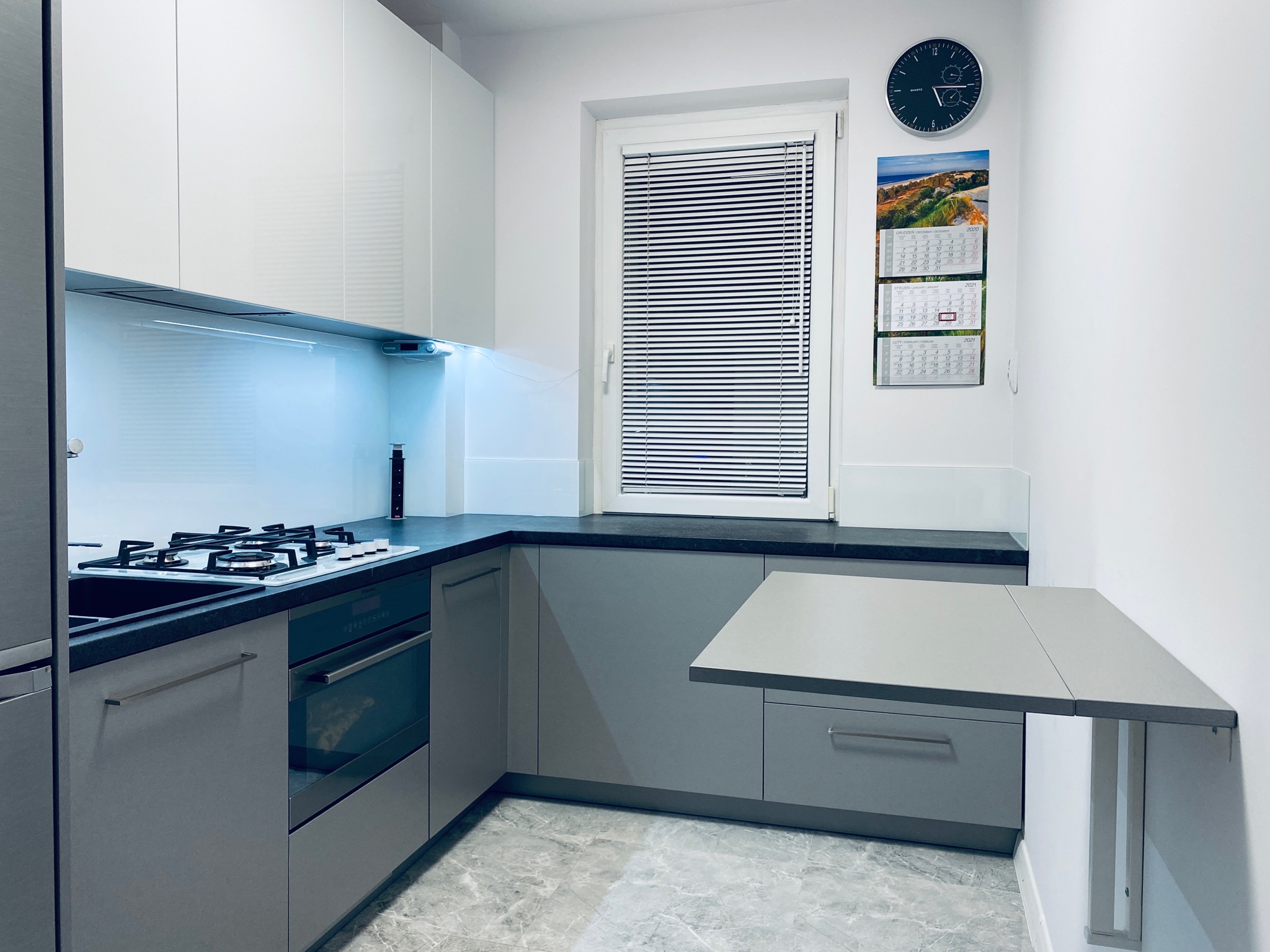 Nowoczesna kuchnia w odcieniach szarości i bieli z blatem roboczym, płytą gazową i piekarnikiem. Widoczny składany blat przymocowany do ściany, zegar i kalendarz na ścianie.