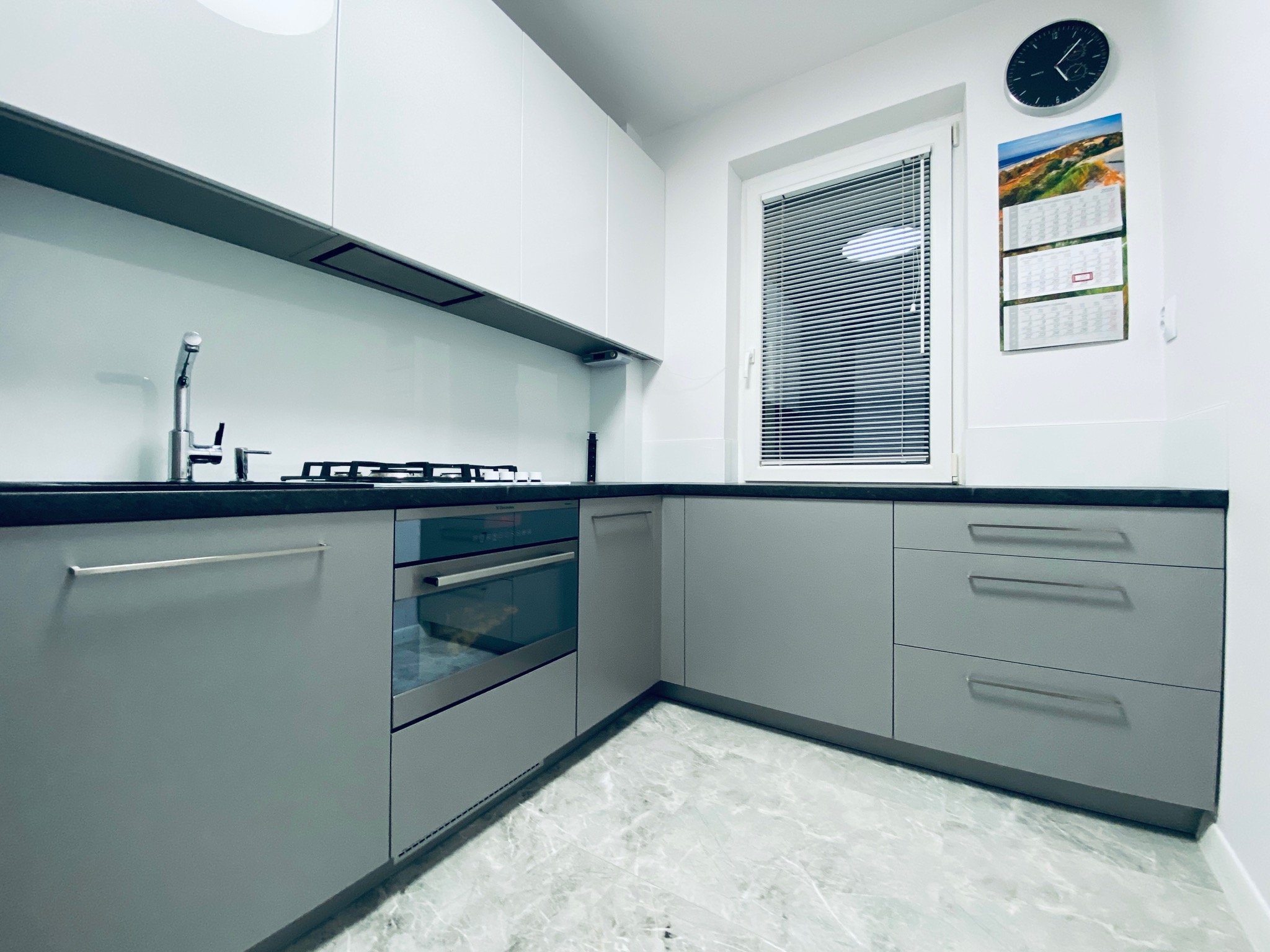 Nowoczesna kuchnia w odcieniach szarości i bieli z czarnym blatem, minimalistycznym okapem i zegarem ściennym, marmurowa podłoga.