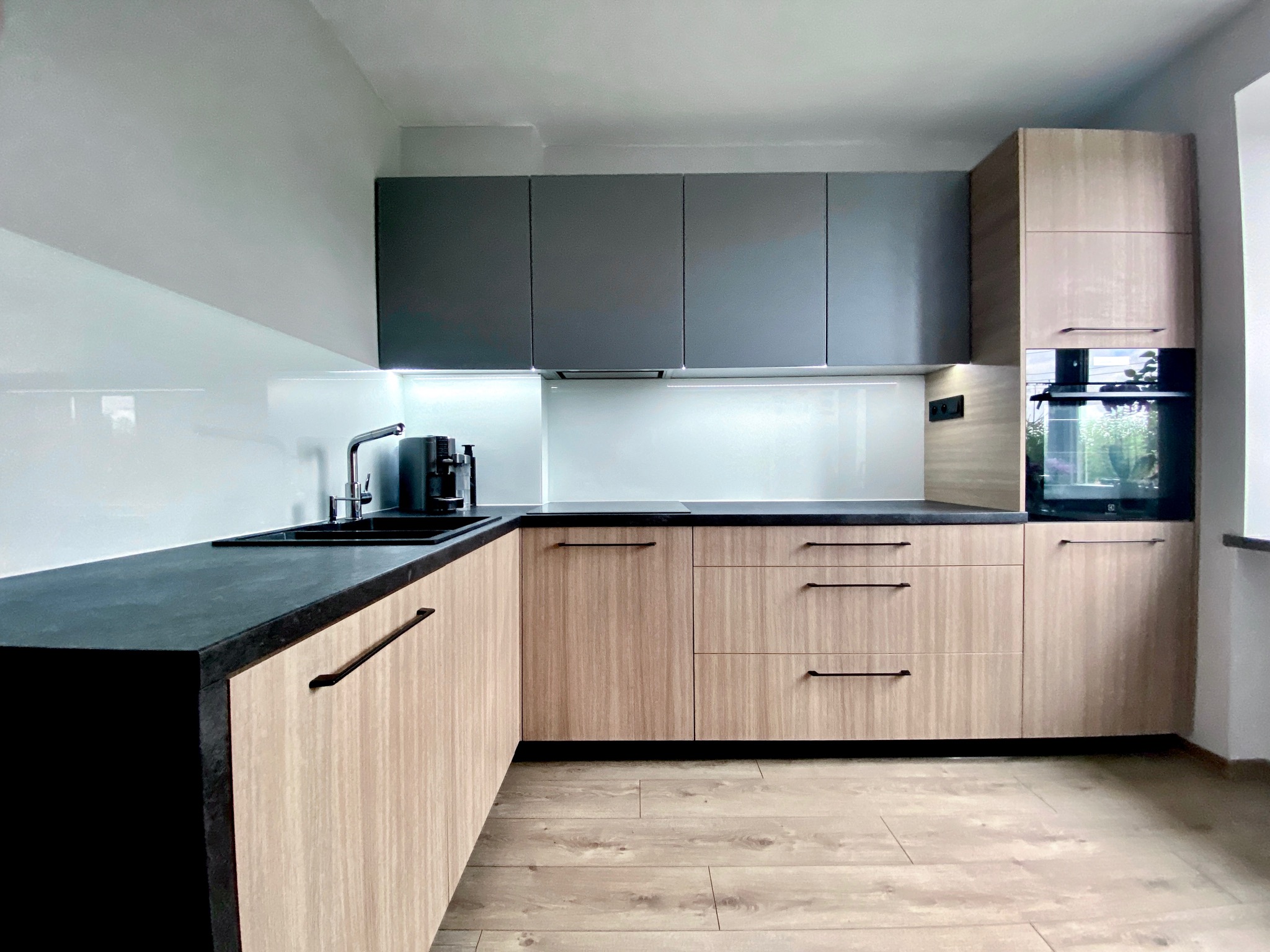 Nowoczesna kuchnia narożna z frontami w odcieniach drewna i szarymi szafkami wiszącymi, czarny blat i minimalistyczne uchwyty, na blacie ekspres do kawy.