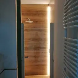 Nowoczesna łazienka z drewnopodobnymi panelami ściennymi i podświetleniem LED, widok z korytarza na wnętrze z grzejnikiem drabinkowym po prawej stronie.