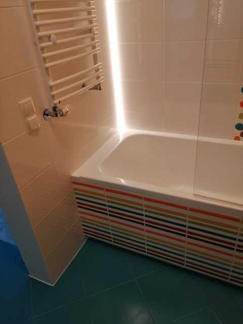 Wykończona łazienka w Sitne z wanną obłożoną płytkami w kolorowe paski, białe płytki na ścianach i grzejnik drabinkowy.