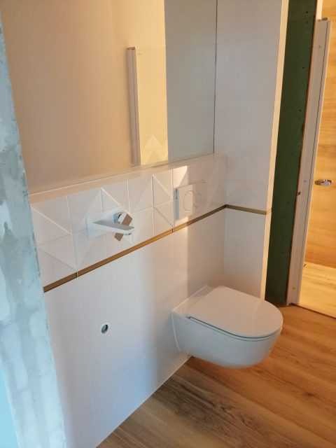 Nowoczesna, podwieszana toaleta z białymi płytkami dekoracyjnymi i złotymi akcentami, zamontowana na drewnianej podłodze w wykończonej łazience.