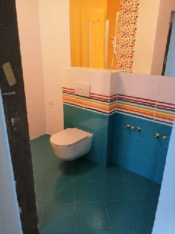 Wykończona łazienka z podwieszaną toaletą, turkusowymi płytkami podłogowymi i ściennymi z poziomym pasem kolorowych pasków, widoczny fragment prysznica z żółtą szybą i mozaiką.