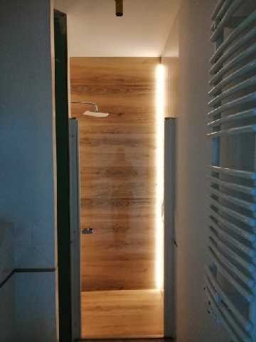 Nowoczesna łazienka z drewnopodobnymi panelami ściennymi i podświetleniem LED, widok z korytarza na wnętrze z grzejnikiem drabinkowym po prawej stronie.