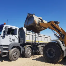 Żółta ładowarka Liebherr wysypuje ziemię do szarej wywrotki MAN TGA 35.440 na piaszczystym terenie pod błękitnym niebem.