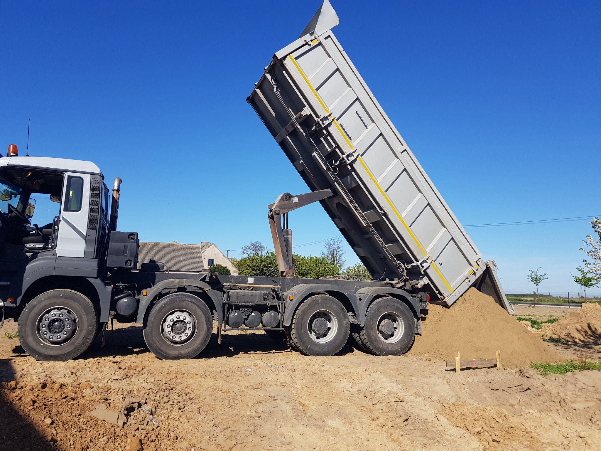 Ciężarówka z podniesioną skrzynią wyładowuje piasek na placu budowy w słoneczny dzień, widoczne drewniane znaczniki w gruncie.