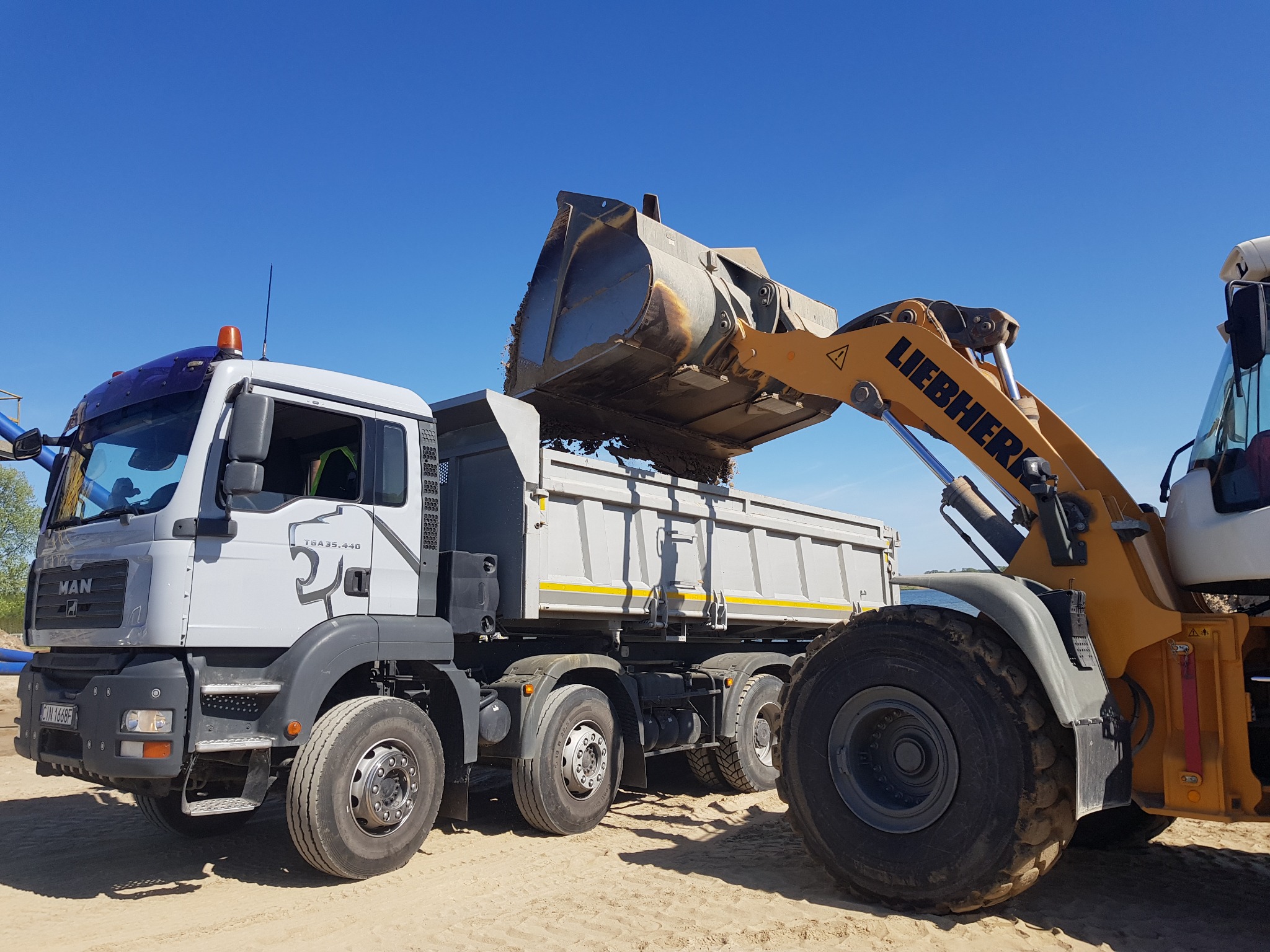 Żółta ładowarka Liebherr wysypuje ziemię do szarej wywrotki MAN TGA 35.440 na piaszczystym terenie pod błękitnym niebem.