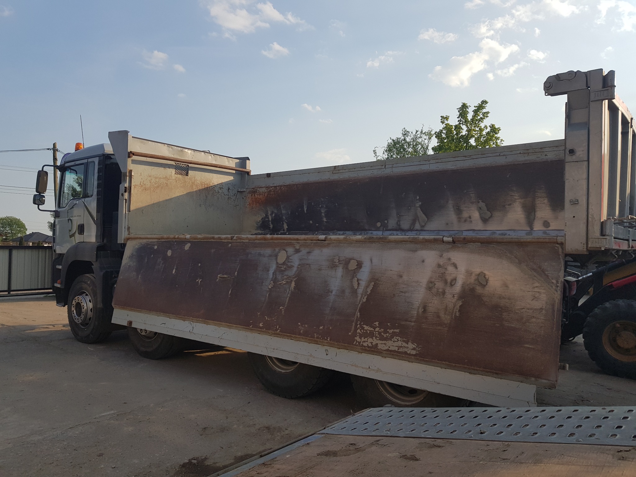 Ciężarówka wywrotka z otwartą klapą, widoczne ślady użytkowania i zabrudzenia na skrzyni ładunkowej, częściowo widoczny pojazd budowlany z tyłu, błękitne niebo w tle.