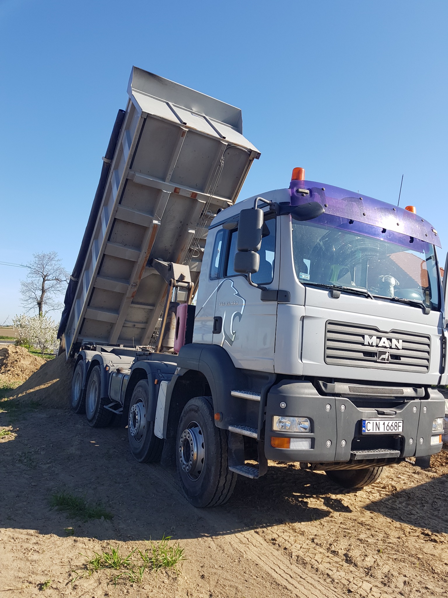 Ciężarówka MAN z podniesioną skrzynią ładunkową wysypuje ziemię na placu budowy w słoneczny dzień. Widoczny numer rejestracyjny CIN 1668F.