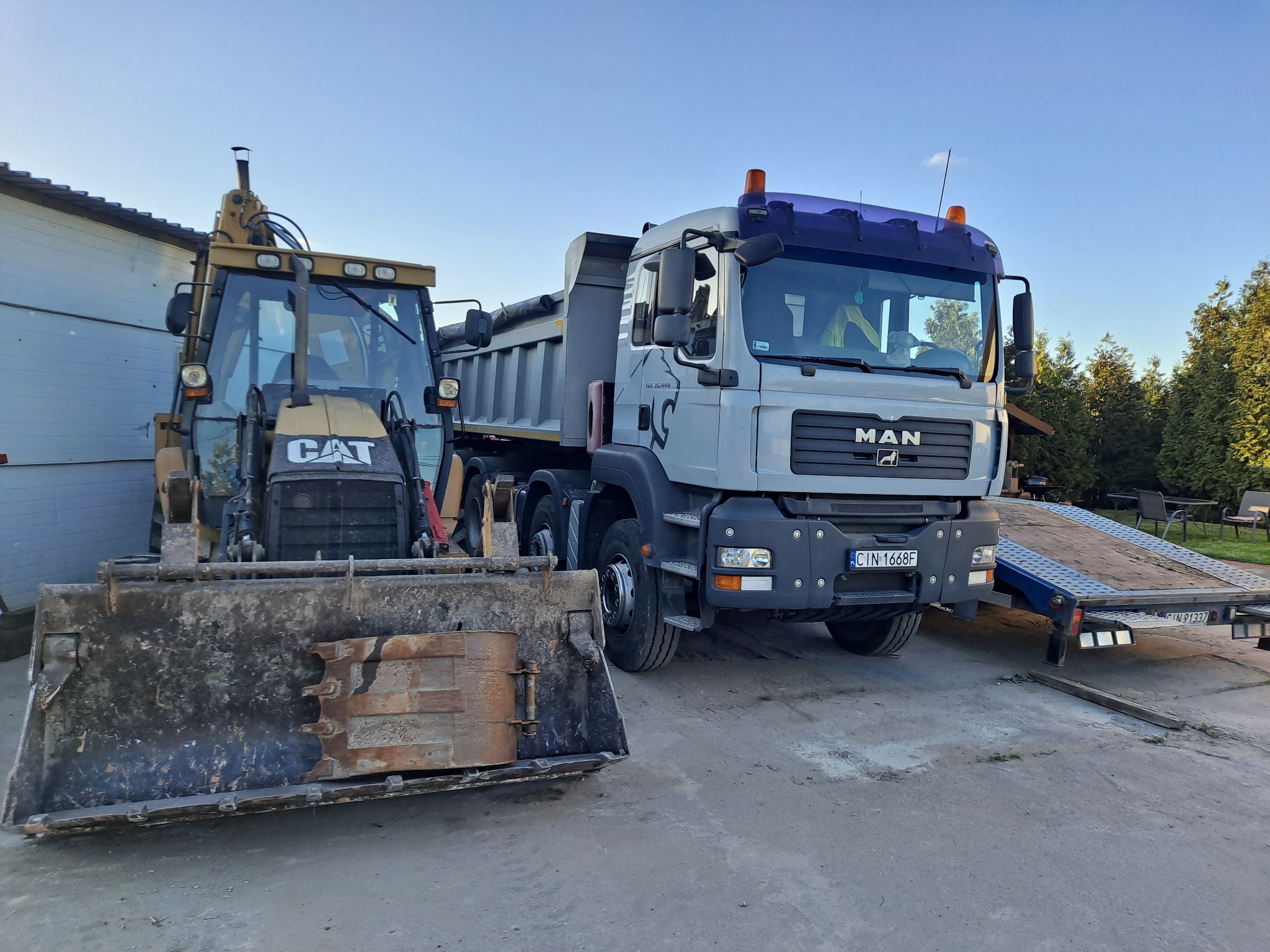 Żółta koparko-ładowarka CAT z brudną łyżką i szara wywrotka MAN z przyczepą na tle budynku i drzew.