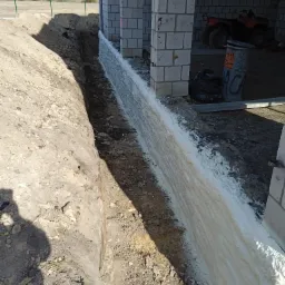 Fundament budynku z bloczków, zabezpieczony białą pianą izolacyjną w wykopie, widoczny ATV w tle budynku.