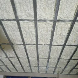 Poddasze z widoczną konstrukcją metalową wypełnioną warstwą białej, spienionej izolacji. Z dołu widoczne okno.