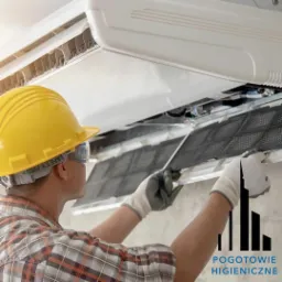 Pracownik w kasku i okularach ochronnych zdejmuje filtr z klimatyzatora, widoczny fragment logo firmy Pogotowie Higieniczne w prawym dolnym rogu.