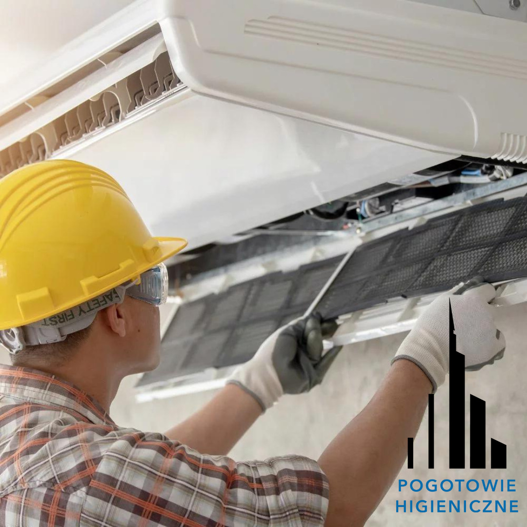 Pracownik w kasku i okularach ochronnych zdejmuje filtr z klimatyzatora, widoczny fragment logo firmy Pogotowie Higieniczne w prawym dolnym rogu.
