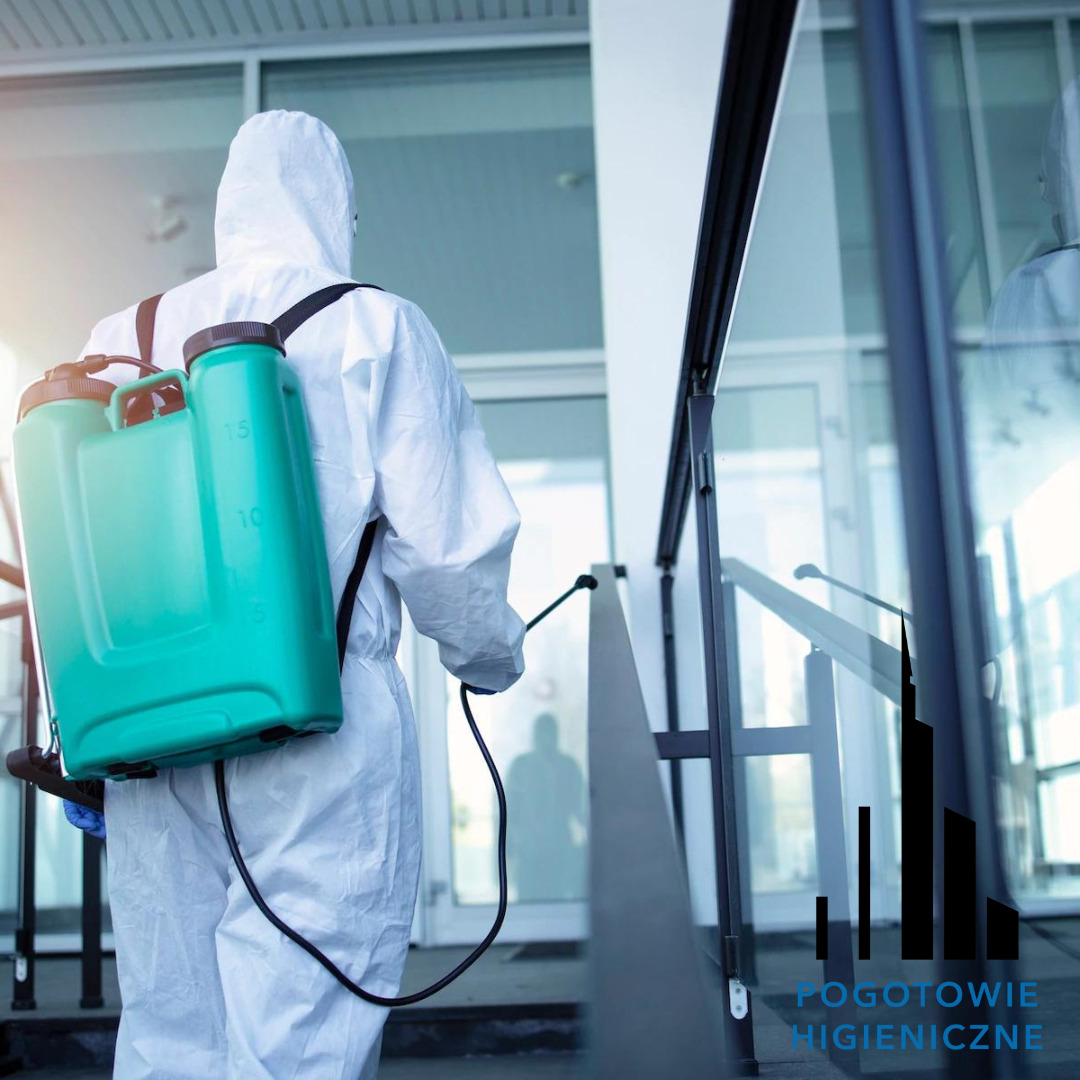 Osoba w kombinezonie ochronnym, z zielonym opryskiwaczem na plecach, dezynfekuje poręcze schodów wejściowych do budynku z przeszklonymi drzwiami.
