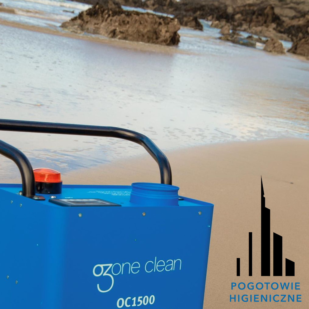 Niebieski generator ozonu Ozone Clean OC1500 z logiem Pogotowie Higieniczne na piaszczystej plaży z widokiem na morze i skały.