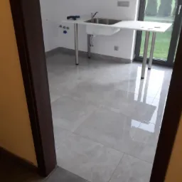 Wnętrze łazienki w trakcie wykańczania, widoczne szare płytki podłogowe z połyskiem, umywalka na metalowych nogach, narzędzia na blacie, okno z widokiem na zieleń na zewnątrz.