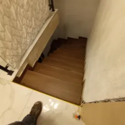 Widok z góry na drewniane schody w trakcie montażu, z widocznymi niedoskonałościami na ścianie i fragmentem narzędzi, a także stopą osoby robiącej zdjęcie na marmurowej podłodze.