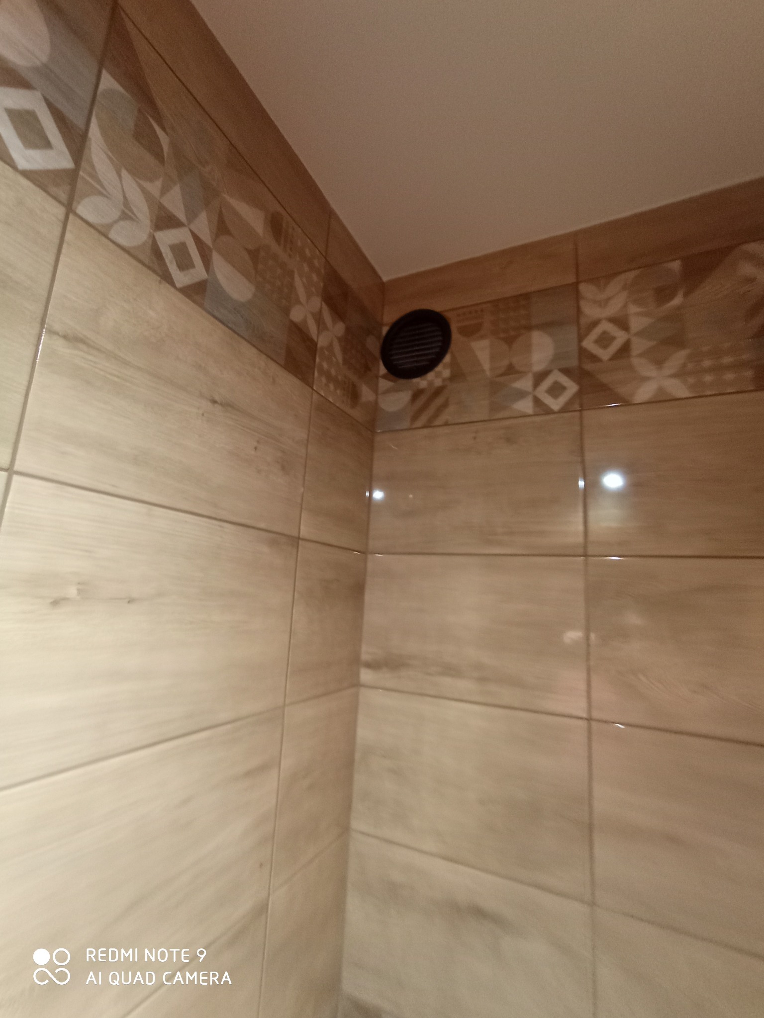 Wykończona ściana w łazience z beżowymi płytkami imitującymi drewno, ozdobny pas płytek z geometrycznym wzorem i okrągła kratka wentylacyjna w rogu.