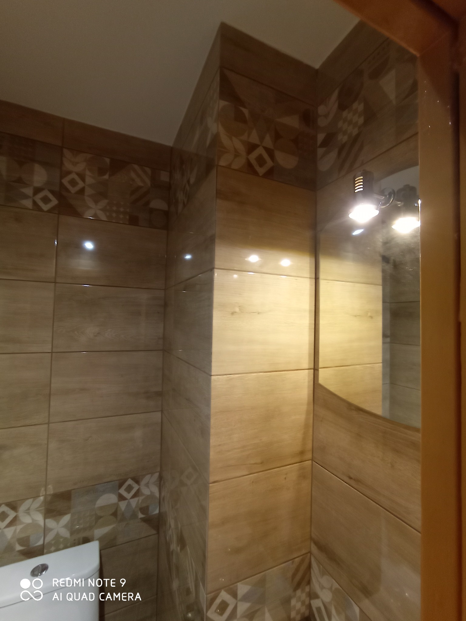 Wykończona łazienka w beżowych płytkach imitujących drewno, z dekoracyjnym pasem płytek z geometrycznym wzorem u góry i dołu, widoczne odbicie oświetlenia w lustrze.