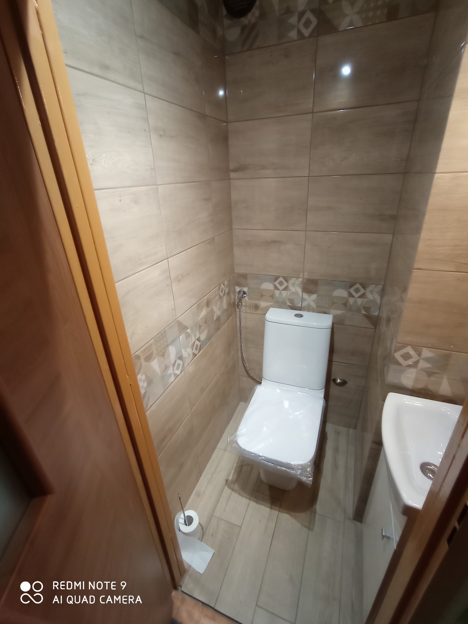 Ukończona łazienka z beżowymi płytkami imitującymi drewno, dekoracyjnym pasem płytek z geometrycznym wzorem, białą toaletą z folią ochronną na desce, umywalką i drewnianą ościeżnicą drzwi.