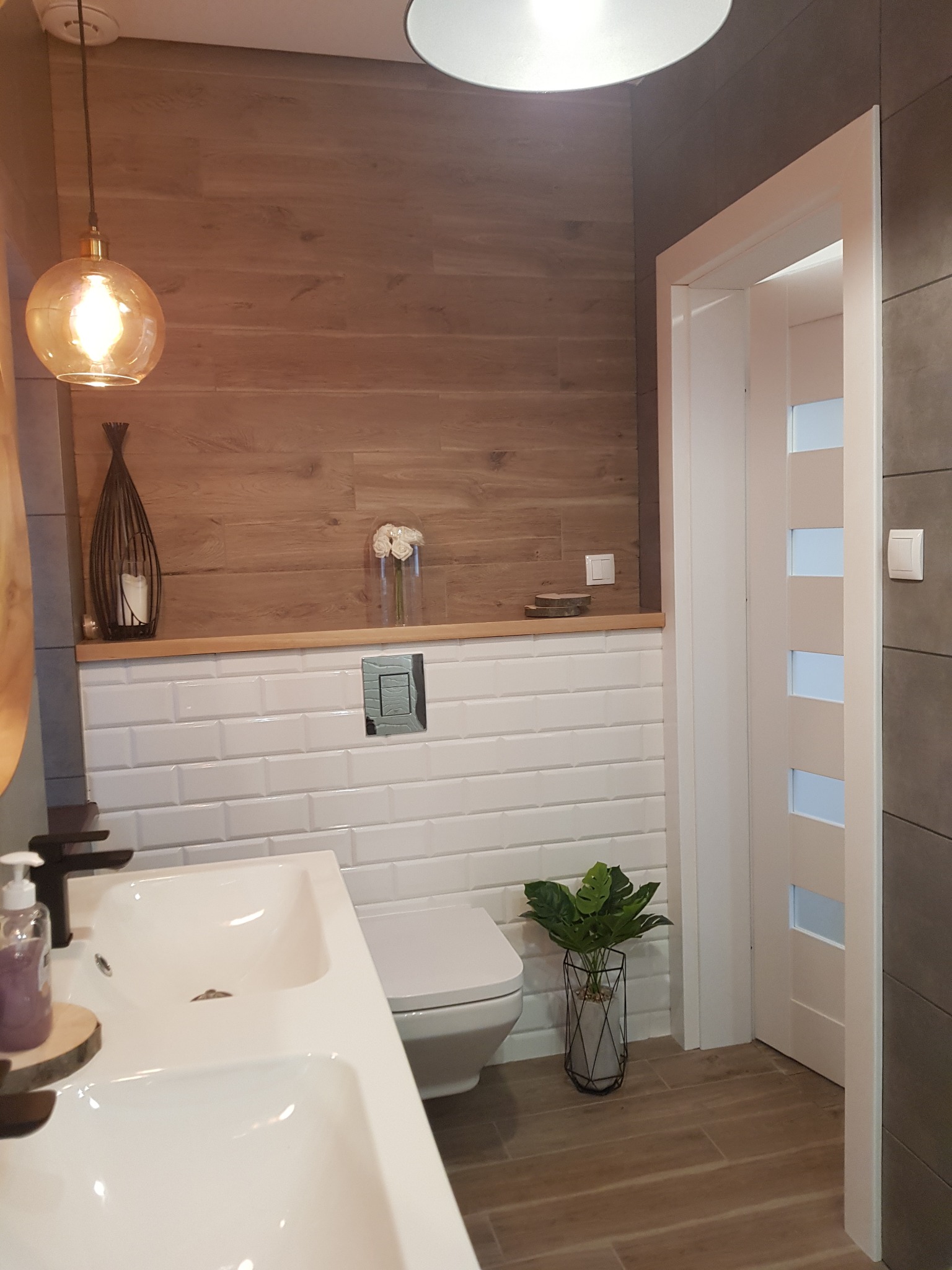 Nowoczesna łazienka z drewnopodobnymi płytkami na ścianie, białą armaturą i dekoracyjną rośliną w metalowym koszu.